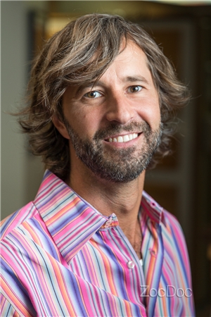 Portrait of a smiling man with shoulder-length brown hair and a beard wearing a pink striped button-down shirt.